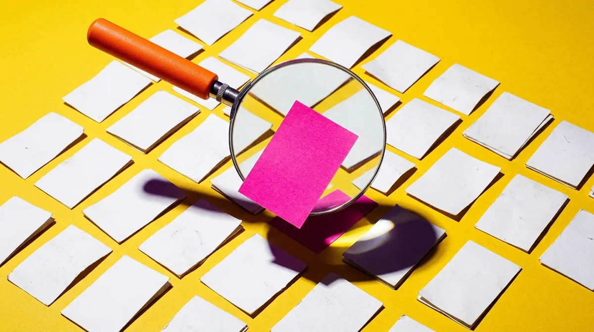 An orange magnifying glass over a grid of white cards, with a single card lifting up and standing out from the rest